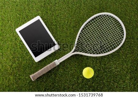 top view of tennis racket and ball near digital tablet with blank screen on green grass, sports betting concept