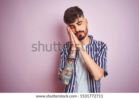 Young man with tattoo wearing striped shirt standing over isolated pink background sleeping tired dreaming and posing with hands together while smiling with closed eyes.