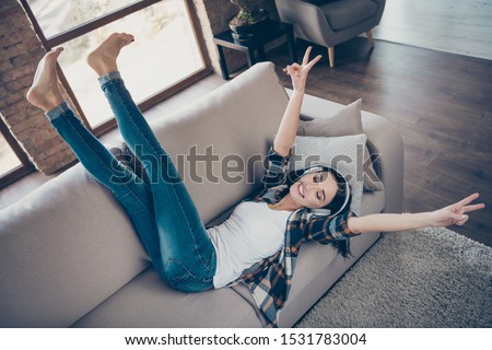 High angle view photo of amazing lady listening favorite radio fm in modern earflaps raising legs up showing v-signs lying comfy sofa wear casual clothes apartment indoors
