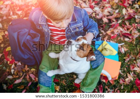 School boy with backpack sitting on ground spangled bright colorful leaves and hugging his dog in the fall autumn park, outdoor. Happy child with puppy friendship, true love, lifestyle concept.
