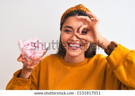 Young beautiful woman holding bowl with marshmallows over isolated white background with happy face smiling doing ok sign with hand on eye looking through fingers