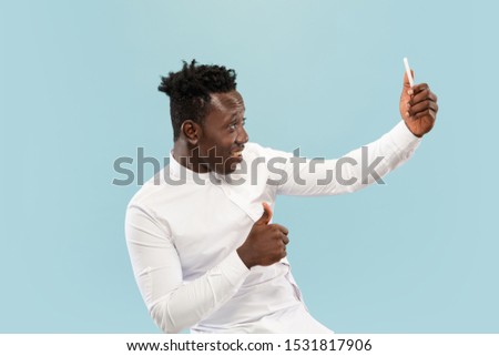 Let's tak a selfie. Young african-american man with smartphone. isolated on blue studio background, facial expression. Beautiful male half-lenght portrait. Concept of human emotions, facial expression