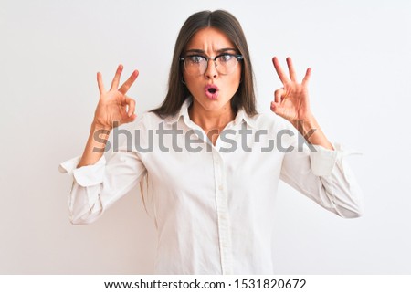 Young beautiful businesswoman wearing glasses standing over isolated white background looking surprised and shocked doing ok approval symbol with fingers. Crazy expression