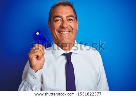 Handsome middle age businessman holding credit card over isolated blue background with a happy face standing and smiling with a confident smile showing teeth