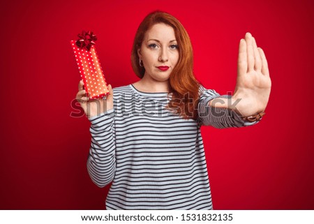 Young beautiful redhead woman holding valentine gift over red isolated background with open hand doing stop sign with serious and confident expression, defense gesture