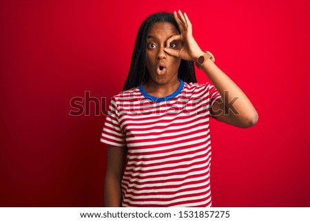 Young african american woman wearing striped t-shirt standing over isolated red background doing ok gesture shocked with surprised face, eye looking through fingers. Unbelieving expression.