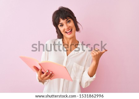 Young beautiful teacher woman reading book standing over isolated pink background pointing and showing with thumb up to the side with happy face smiling