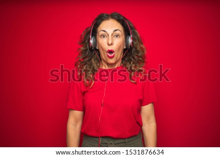 Middle age senior woman wearing headphones listening to music over red isolated background afraid and shocked with surprise expression, fear and excited face.
