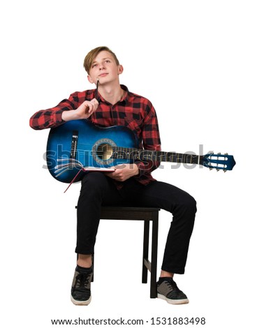 Full length portrait of talented boy teenager with his trendy acoustic guitar seated on chair thinking to compose a new song while holding a pencil and notebook isolated over white background.
