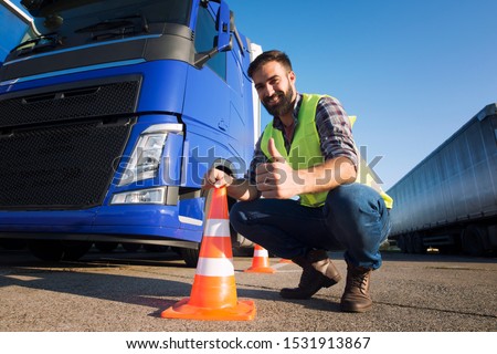 Shot of bearded man learning how to drive truck at driving school. Truck driver candidate training for driving license. Standing by the traffic cone in reflective vest. Truck in the background. Royalty-Free Stock Photo #1531913867