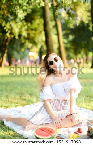Young caucasian nice woman in sunglasses having picnic on plaid and sitting in park with fruits. Concept of resting in open air, leisure time and summer season