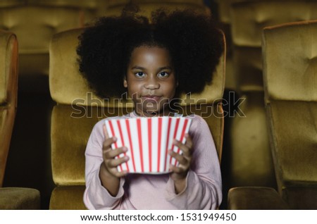 Cute little African American black girl eating popcorn and enjoying a movie at the cinema theater, looking at camera. Concept entertainment and enjoyment.