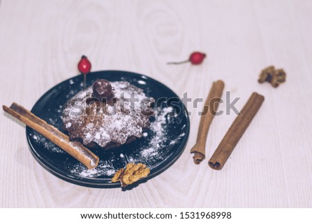 Cupcake with berry and icing sugar in a black plate and cinnamon sticks and walnuts on a light table background. Selective focus.
