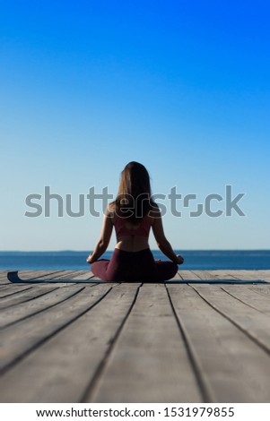 silhouette backwards of a slender young woman with long hair sitting in lotus yoga pose sukhasana on wooden platform at the sea coast in summer sunset 