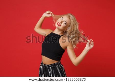 Smiling pretty beautiful young woman girl in black clothes posing isolated on bright red wall background, studio portrait. People sincere emotions lifestyle concept. Mock up copy space. Holding hair