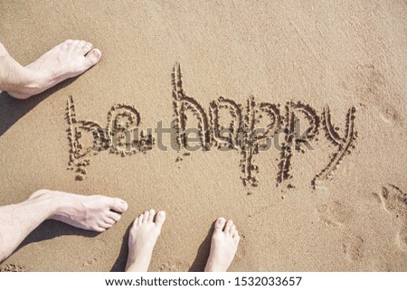 Legs of couple stands near words be HAPPY drawn on the sand of sandy beach. Vacation concept.