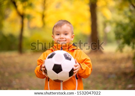 Portrait of toddler boy with soccer ball in autumn park. Smiling little child with funny face in orange jacket holding football ball. Healthy childhood, sports and outdoor activities concept