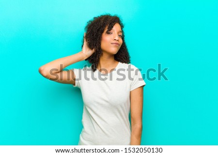 young black woman smiling, looking curiously to the side, trying to listen to gossip or overhearing a secret against blue wall