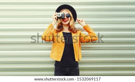 Stylish smiling woman with retro camera taking picture wearing yellow jacket, black round hat on metal wall background