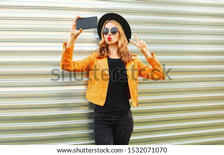 Stylish woman taking selfie picture by smartphone wearing yellow jacket, black round hat on metal wall background