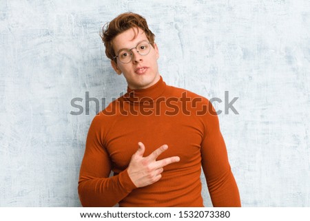 young red head man feeling happy, positive and successful, with hand making v shape over chest, showing victory or peace against grunge wall