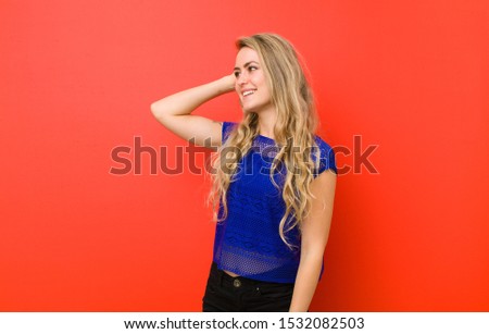young blonde woman smiling cheerfully and casually, taking hand to head with a positive, happy and confident look against red wall