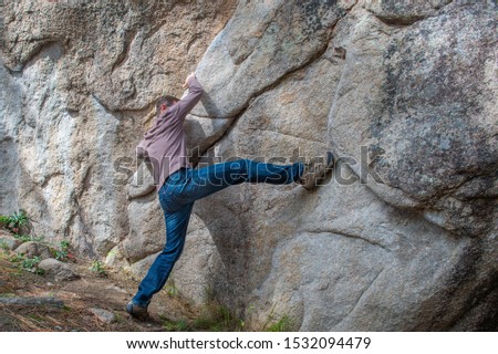 One giant step: A man, casually dressed and wearing hiking boots, secures a finger hold in a fissure on a granite rock wall and takes one awkward, large step to begin his climb.