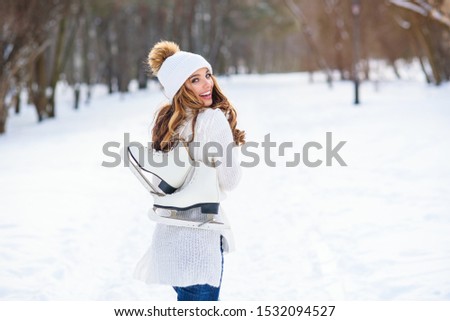 Beautiful woman weared in white sweater and hat with ice skates on the back walks in winter snowy park.