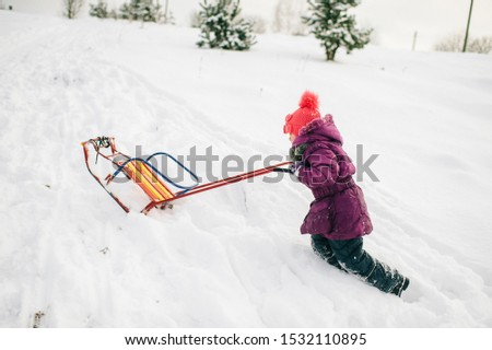 Little girl pushing sled up the snowy slide in witer day outdoors. Winter entartainment.