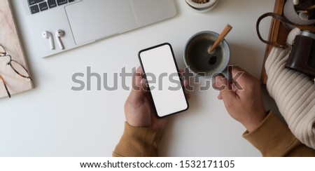 Top view of man looking at blank screen smartphone while holding coffee cup in cozy workplace