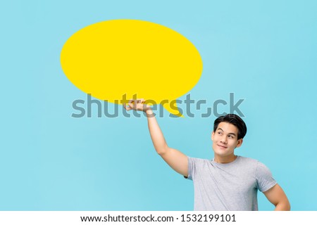 Handsome smiling Asian man thinking and looking at empty speech bubble isolated on light blue background