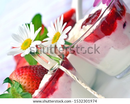  close up of two glasses with natural yoghurt and silver fork, fresh, fruity strawberries,  served on a wooden white table with tablecloth , decorated with white daisies.