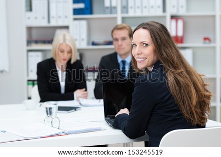 Businesswoman in a meeting sitting with coworkers at a table in the office turning in her chair to smile at the camera