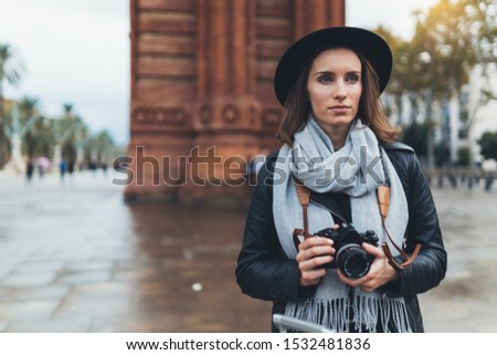 Photographer tourist with retro photo camera. Young girl in hat travels in Triumphal arch Barcelona. Holiday concept street in europe city. Traveler hipster shooting architecture, copy space mockup