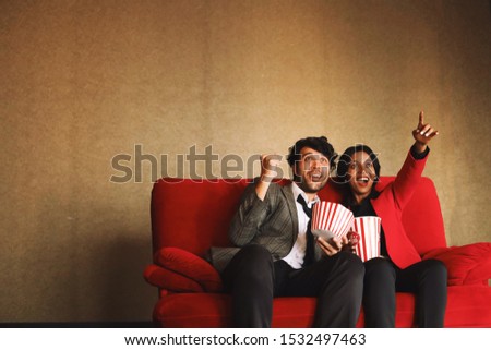 Two exciting people sitting on red sofa and holding popcorn buckets.