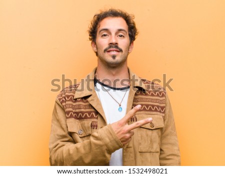 young handsome man feeling happy, positive and successful, with hand making v shape over chest, showing victory or peace against orange wall
