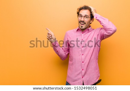 young handsome man laughing, looking happy, positive and surprised, realizing a great idea pointing to lateral copy space against orange wall