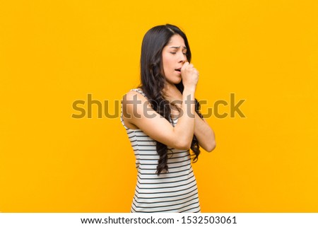 young pretty latin woman feeling ill with a sore throat and flu symptoms, coughing with mouth covered against orange wall