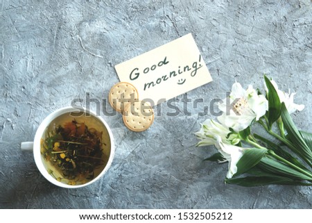 Beautiful white alstroemeria flowers  with a cup of herbal tea and a Good Morning message on pastel paper with a biscuits. Top view, flat lay. Textured grey background. 