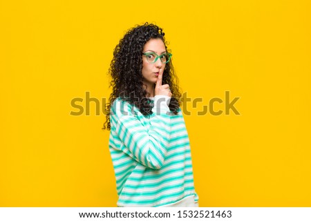 young pretty woman asking for silence and quiet, gesturing with finger in front of mouth, saying shh or keeping a secret against orange wall