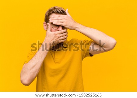 young blonde man covering face with both hands saying no to the camera! refusing pictures or forbidding photos against orange wall