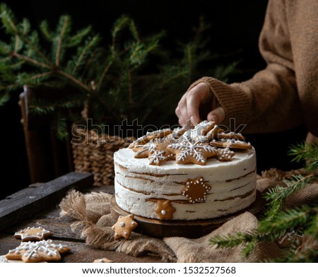 woman decorates homemade round multilayered cake with white cream and gingerbread snowflakes shaped cookies on rustic wooden table with sackcloth, fir tree branches 