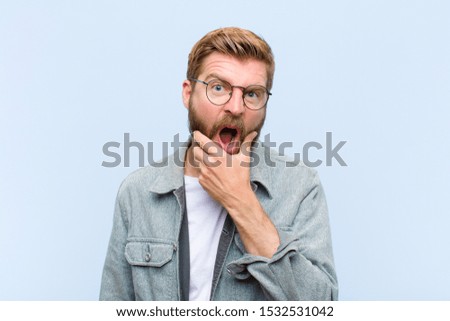 young blonde adult man with mouth and eyes wide open and hand on chin, feeling unpleasantly shocked, saying what or wow