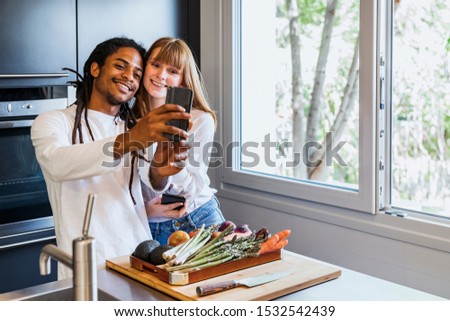 Stock photo of a Multiethnic couple doing a selfie with the mobile in the kitchen next to the window and with a box of vegetables, a cutting table and a knife in front. Life style