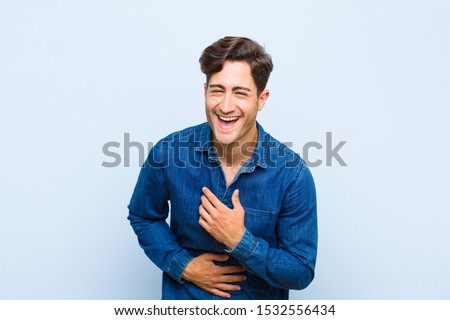 young handsome man laughing out loud at some hilarious joke, feeling happy and cheerful, having fun against blue background