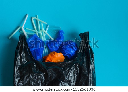 Black plastic bag with variety elements of plastic in it, against a blue background. Representation of plastic pollution Royalty-Free Stock Photo #1532561036