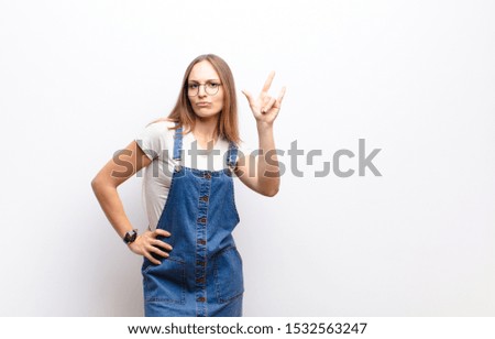 young pretty woman feeling happy, fun, confident, positive and rebellious, making rock or heavy metal sign with hand against white wall