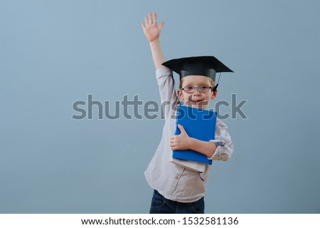Cute smiling little ginger boy in glasses, wearing student hat holds stack of books in hands over blue background. Half length. He's eagerly raising his arm in a fashion to answer teachers question.