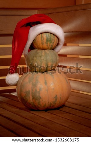 A pyramid of three pumpkins on a wooden surface is decorated with a Santa Claus hat. Through the walls of wooden slats light breaks through with beautiful rays. Festive warm picture for autumn, winter