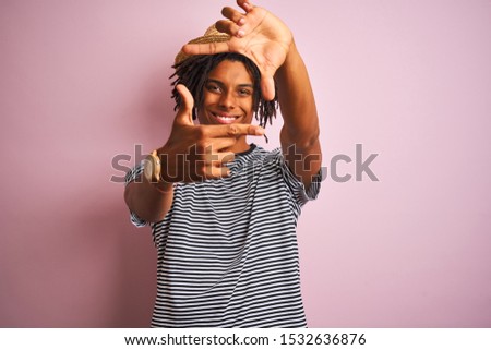 Afro american man with dreadlocks wearing navy t-shirt and hat over isolated pink background smiling making frame with hands and fingers with happy face. Creativity and photography concept.